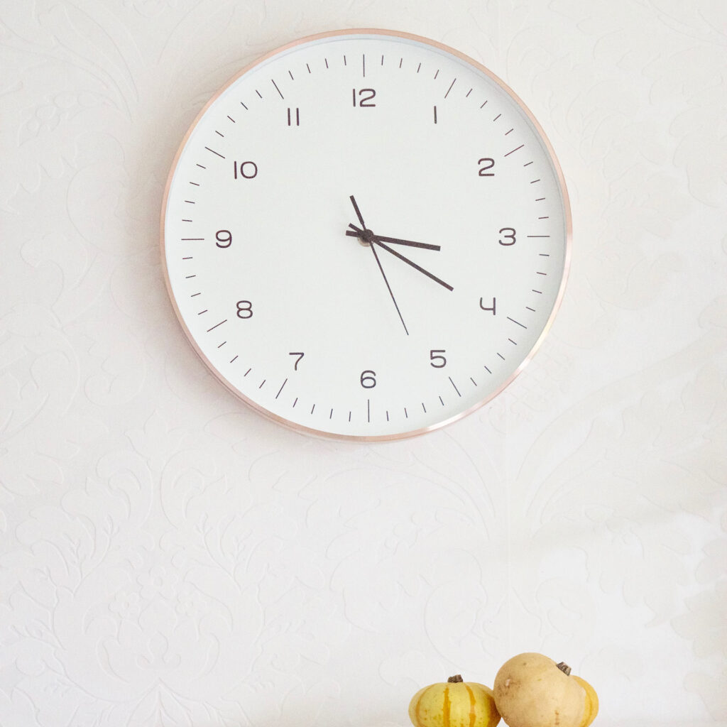 A white wall clock showing 3:17 with two small pumpkins below it.